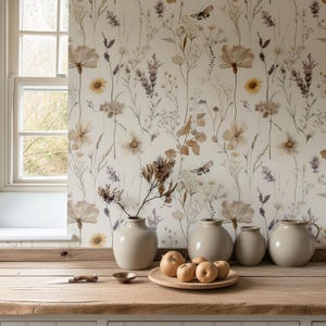 May include: A neutral-toned interior scene featuring a wooden table with a selection of decorative items. The table holds a wooden plate with apples, small ceramic bowls, and a collection of beige vases. The backdrop is a floral wallpaper with a window.