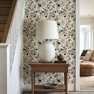 May include: A wooden side table with a white lamp, a small brown vase, and a decorative bowl. The wall behind the table is covered in floral wallpaper with black, brown, and cream-colored flowers. A staircase and doorway are visible.
