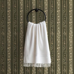 May include: A white, fringed towel hangs from a black metal ring. The towel is folded over the ring, and the background is a green and beige striped wallpaper with floral accents. The towel has a textured appearance.