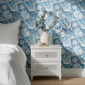 May include: A white nightstand with two drawers featuring woven cane fronts. A white vase with dried stems and a small bowl sit on top. The background is a blue and white oyster shell patterned wallpaper.
