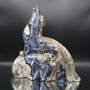 Puede incluir: Escultura de piedra de un lobo aullando. El lobo está representado en posición sentada, con la cabeza levantada y la boca abierta. La escultura está hecha de piedra con colores grises, negros y beige. Está sobre una superficie reflectante.