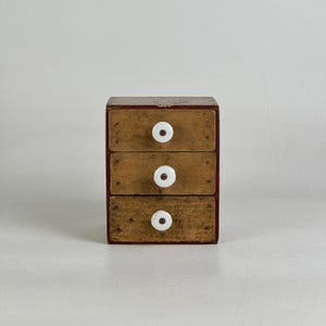May include: A small wooden storage chest featuring three stacked drawers with a weathered, light brown finish. Each drawer is equipped with a round, white ceramic knob centered on the front. The outer frame of the chest is painted a dark red.