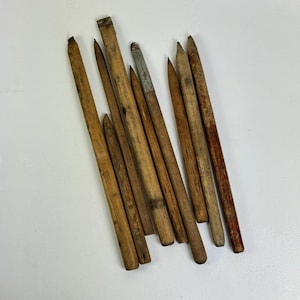 May include: A collection of vintage wooden pencils of varying lengths and shades of brown. Some pencils have a sharpened point, while others have a blunt end. One pencil has a metal band with faded text.