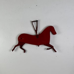 May include: A flat, red-painted metal horse figure in a galloping pose. The horse features a triangular metal attachment extending upward from the center of its back, with articulated legs and a small circular hole visible on the rear flank.
