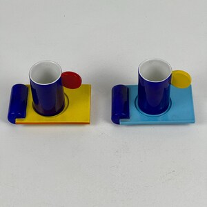May include: Two blue and white espresso cups with colorful geometric designs on the saucers. One saucer is yellow, orange, and blue. The other saucer is blue and light blue.