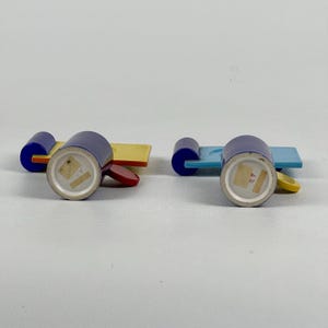 May include: Two blue ceramic cups with yellow, red, and blue geometric designs. The cups are on matching saucers with a yellow, red, and blue geometric design. The cups have a white interior and a white bottom with a blue mark.