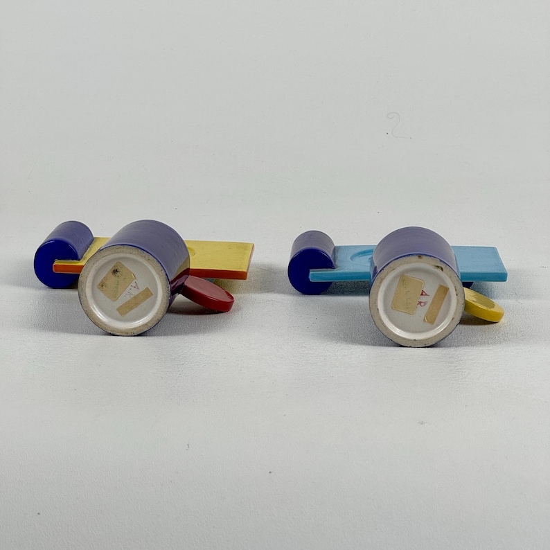 May include: Two blue ceramic cups with yellow, red, and blue geometric designs. The cups are on matching saucers with a yellow, red, and blue geometric design. The cups have a white interior and a white bottom with a blue mark.