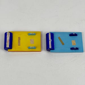 May include: Two small wooden platforms with blue, yellow, and light blue paint. Each platform has a blue cylinder on one end and two light blue circles on the other end.