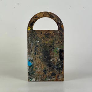 May include: A rectangular wooden artist's palette with a handle. The palette is heavily stained with dried paint in various colors, including blue, yellow, green, and black. The palette has a semi-circular handle at the top.