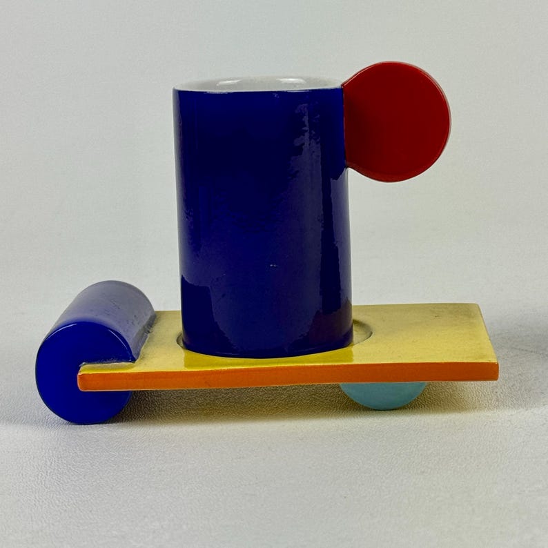 May include: A blue ceramic cup with a red handle sits on a yellow tray with orange and blue accents. The tray has a rounded edge and a blue wheel on one side and a light blue wheel on the other.