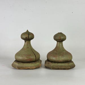 May include: Two weathered, sage green wooden finials with a bulbous, onion-shaped top and a wide, octagonal base. The wood shows visible grain, surface scratches, and a vertical crack on the left piece, indicating a distressed, aged finish.