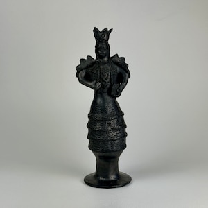 May include: A dark gray ceramic figurine of a person wearing a crown, ornate shoulder pieces, and a layered skirt. The figure holds a small book and stands on a round base. The statue has a detailed, textured surface.
