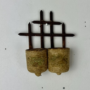 May include: A wall-mounted object featuring two cylindrical, textured, light-tan containers suspended from a grid of four dark, rusted metal screws. The screws are arranged in a cross-hatch pattern, with two vertical screws extending into each container.