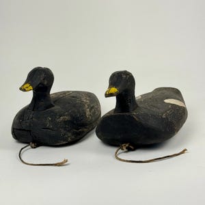 May include: Two vintage wooden duck decoys with black paint and yellow beaks. The decoys have a string attached to their heads.