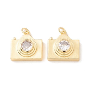 May include: Two gold-colored camera charms with a clear, round gem in the center of the lens. The charms have a loop at the top for attaching to a chain or bracelet. The camera charms are a jewelry-making supply.