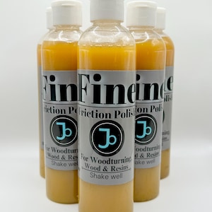 May include: Four amber glass bottles with white lids and labels. The labels are gray with black text that reads "FinFine" and "Friction Polishion Polish". Each bottle has a blue and black logo with the letter "J" inside a circle. The text on the labels reads "For Woodturning, Wood & Resins, Shake well".