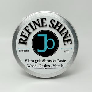May include: A silver tin with a white label that says "REFINE SHINE" in black lettering. The label also features a black circle with a blue "J" inside. The text "Non-Toxic" and "4oz" are printed on the label. The label also says "Micro-grit Abrasive Paste Wood - Resins - Metals".