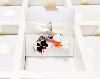 May include: A silver charm featuring a white dog with an orange scarf and a dangling rectangular pendant shaped like a playing card. The card displays a black silhouette of the dog, a red diamond suit, and the text Flying Ace in black script.