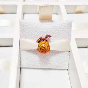 May include: A small, rounded glass bead charm featuring a stylized, dark reddish-brown fox figure perched atop a vibrant, golden-orange floral pattern. The bead has a smooth, glossy finish and is secured by a horizontal cream-colored ribbon.