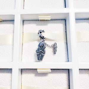 May include: A silver charm featuring an Egyptian pharaoh and a mummy. The pharaoh charm is detailed with a headdress and intricate carvings. The mummy charm is connected by a chain. The charm is displayed in a white box.