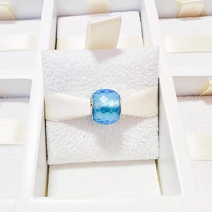 May include: A faceted, light blue bead charm is the focal point, set against a white, textured background. A cream-colored ribbon is wrapped around the bead. The charm is displayed in a white jewelry box, suggesting a gift or presentation setting.