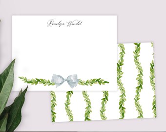 May include: A white stationery set with a green leafy border and a blue bow. The stationery is personalized with the name "Penelope Winslet". The envelope has a repeating pattern of green leafy garlands.
