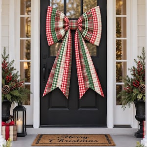 May include: A large decorative bow featuring a red, green, and white plaid pattern attached to a black door. The bow has two wide loops and two long, pointed tails. Below the door, a rectangular brown doormat displays the text Merry Christmas.