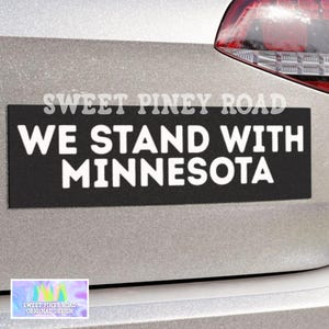 Puede incluir: Una pegatina rectangular negra con el texto blanco "WE STAND WITH MINNESOTA". La pegatina está adherida a un vehículo plateado. El texto "SWEET PINEY ROAD" está encima del texto principal.