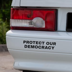 May include: White car bumper with a red and white tail light. The bumper has the text "PROTECT OUR DEMOCRACY" in black, bold letters.