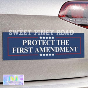 May include: A blue rectangular bumper sticker with the text "PROTECT THE FIRST AMENDMENT" in white, framed by a red border and stars. The sticker is affixed to a car.