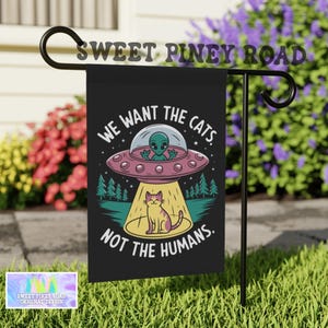 Puede incluir: Bandera de jardín negra con el texto "WE WANT THE CATS NOT THE HUMANS". La bandera presenta un extraterrestre de dibujos animados en un OVNI que proyecta un gato. La bandera también tiene el texto "SWEET PINEY ROAD".
