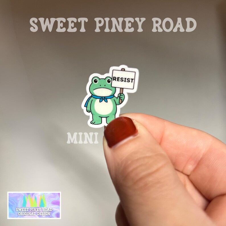 Puede incluir: Una pegatina de rana verde con una bufanda azul y un letrero que dice "RESIST". La pegatina la sostiene una persona. Tambi&eacute;n se ve el texto "SWEET PINEY ROAD" y "MINI".