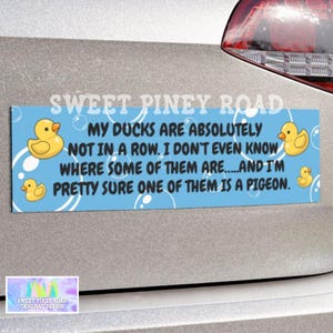 May include: A blue bumper sticker with the text "MY DUCKS ARE ABSOLUTELY NOT IN A ROW...AND I'M PRETTY SURE ONE OF THEM IS A PIGEON." Features yellow rubber duck illustrations and bubble graphics. The sticker is from Sweet Piney Road.
