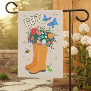Peut inclure: Un drapeau de jardin avec un fond brun clair représentant une botte de pluie jaune remplie de fleurs colorées. La botte est entourée de papillons bleus, d'une abeille jaune et d'une chenille verte. Le texte "FDT" est écrit en lettres jaunes en haut du drapeau.