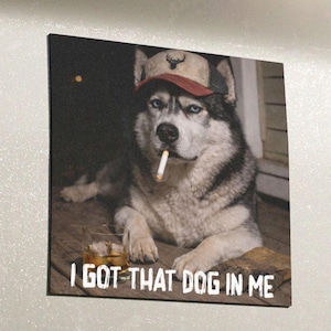 May include: A humorous print featuring a husky dog wearing a hat and smoking a cigarette. The dog is lying down with a glass of whiskey. The text "I GOT THAT DOG IN ME" is printed at the bottom.