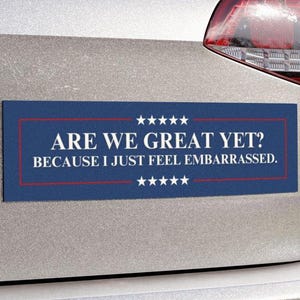 May include: A rectangular bumper sticker with the text "ARE WE GREAT YET? BECAUSE I JUST FEEL EMBARRASSED." The sticker has a blue background with white stars and red borders and text. It is affixed to a silver vehicle.