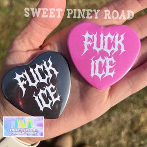 Puede incluir: Dos botones en forma de corazón. Uno es negro con texto blanco que dice "FUCK ICE". El otro es rosa con el mismo texto blanco. Los botones se sostienen en una mano. La parte superior de la imagen dice "SWEET PINEY ROAD".