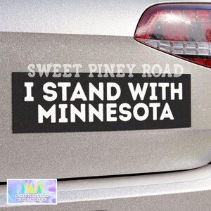 Puede incluir: Una pegatina rectangular negra para parachoques con el texto "I STAND WITH MINNESOTA" en blanco, adherida a la parte trasera de un vehículo plateado. La pegatina también tiene el texto "SWEET PINEY ROAD" en la parte superior.