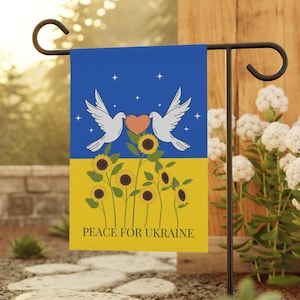 Op de afbeelding: Een blauwe en gele tuin vlag met twee witte duiven die een hart en zonnebloemen vasthouden. De tekst "PEACE FOR UKRAINE" is op de vlag gedrukt.