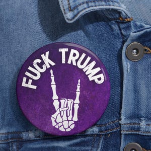 Puede incluir: Un botón redondo morado con el texto blanco "FUCK TRUMP" y un símbolo de mano de esqueleto blanco. El botón está prendido a una chaqueta vaquera azul.