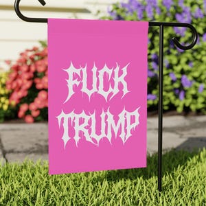 Könnte beinhalten: Eine leuchtend pinkfarbene Gartenflagge mit dem weißen Text "FUCK TRUMP" in gotischer Schrift. Die Flagge ist auf einem schwarzen Metallständer montiert, mit einem Hintergrund aus grünem Gras und bunten Blumen.