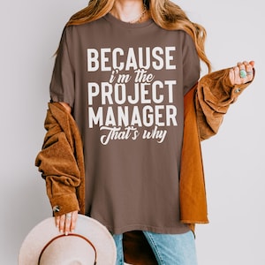 May include: A brown t-shirt with the text "BECAUSE I'm the PROJECT MANAGER That's why" in white. The shirt is worn by a person with long blonde hair, wearing a brown jacket and blue jeans.