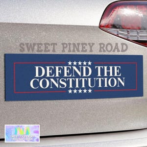 May include: Blue and red bumper sticker with white text that reads "DEFEND THE CONSTITUTION" with five stars above and below the text. The sticker is on a silver car.