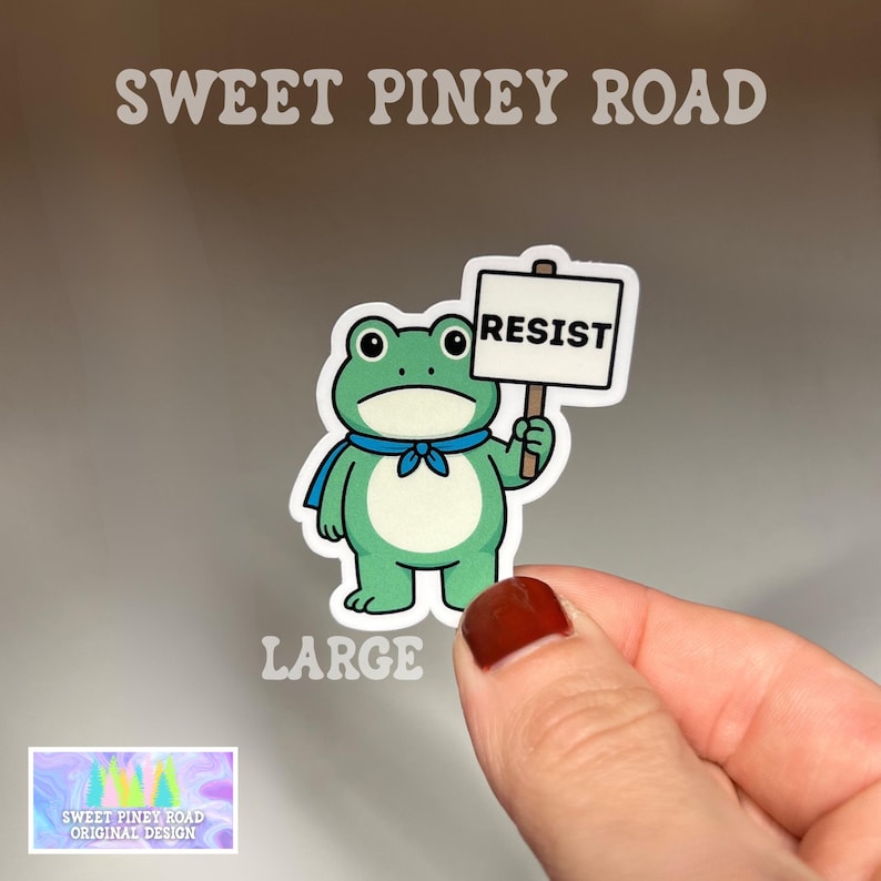 Puede incluir: Una pegatina de rana verde con una bufanda azul, sosteniendo un cartel que dice "RESIST". La pegatina est&aacute; etiquetada como "LARGE" y presenta el texto "SWEET PINEY ROAD ORIGINAL DESIGN". La rana tiene ojos grandes y redondos.
