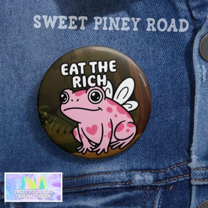 May include: A round button with a pink frog illustration with heart-shaped spots and white wings. The text "EAT THE RICH" is above the frog. The button is set against a denim background, with the text "SWEET PINEY ROAD" at the top.