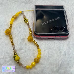 May include: A yellow beaded phone charm with a clear phone case. The phone case has the text "SWEET PINEY ROAD" printed on it.