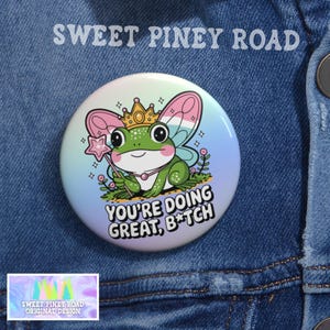 Könnte beinhalten: Ein runder Button mit einer Cartoon-Froschfigur, die eine Krone und Feenflügel trägt und einen Sternenstab hält. Der Button trägt den Text "YOU'RE DOING GREAT, B*TCH" und "SWEET PINEY ROAD" in weißer Schrift auf einem Jeanshintergrund.