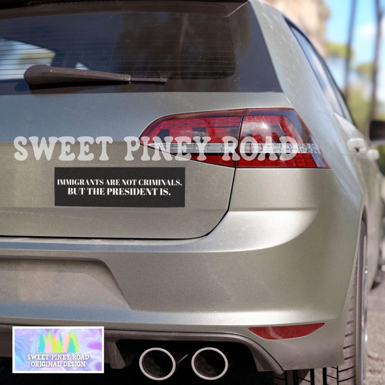 May include: A silver car with a black bumper sticker that reads "IMMIGRANTS ARE NOT CRIMINALS, BUT THE PRESIDENT IS." The text "SWEET PINEY ROAD" is above the bumper sticker. A smaller sticker with the text "SWEET PINEY ROAD ORIGINAL DESIGN" is below.