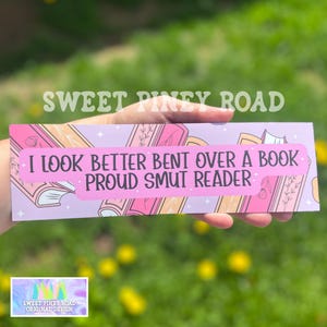 Könnte beinhalten: Rosa-lilafarbenes Lesezeichen mit einem Stapel Bücher und dem Text "I look better bent over a book. Proud smut reader."