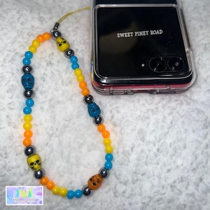 Puede incluir: Un charm de teléfono colorido con una cadena de cuentas y dijes de calavera en amarillo, naranja, azul y gris. El charm está unido a una funda de teléfono transparente con el texto "SWEET PINEY ROAD" impreso en la parte posterior.